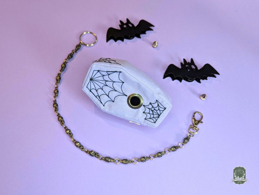 lilac coffin shaped poop bag holder with black embroidered spiderwebs on a pastel pink background