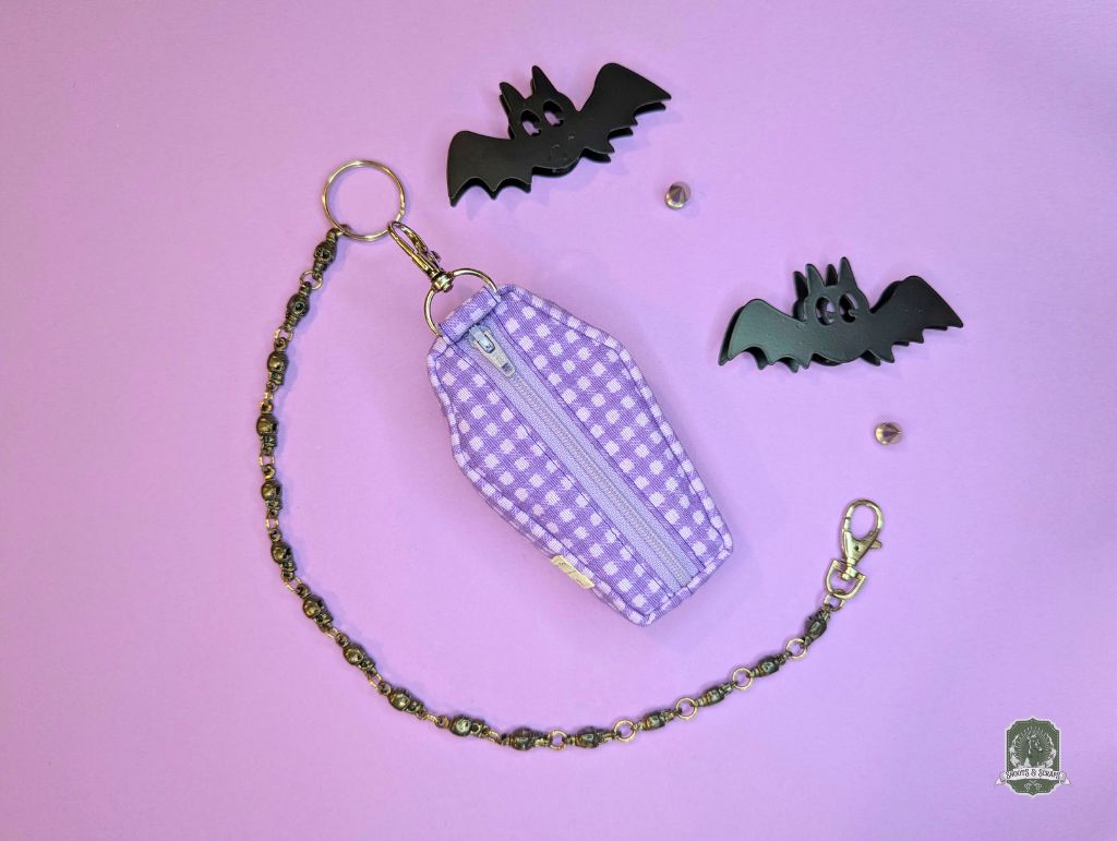 Purple checkered coffin-shaped poop bag holder dispenser pouch with chain on a pink background with black bat decorations.