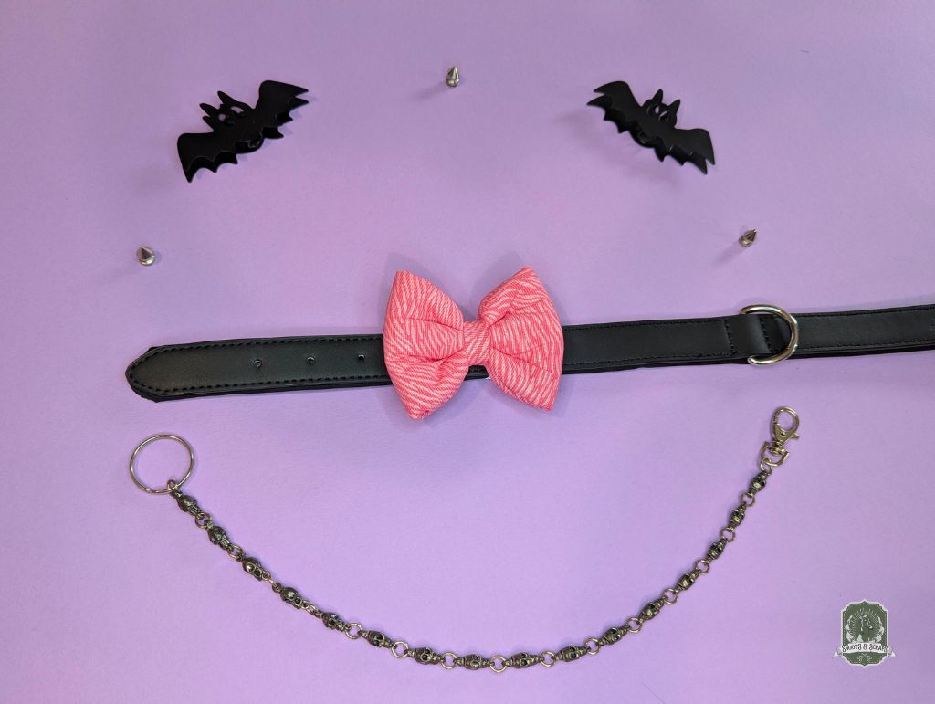 Pink zebra print dog bow tie displayed on a black leather collar with a pastel pink background and decorative chain.