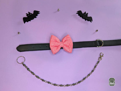 Pink zebra print dog bow tie displayed on a black leather collar with a pastel pink background and decorative chain.
