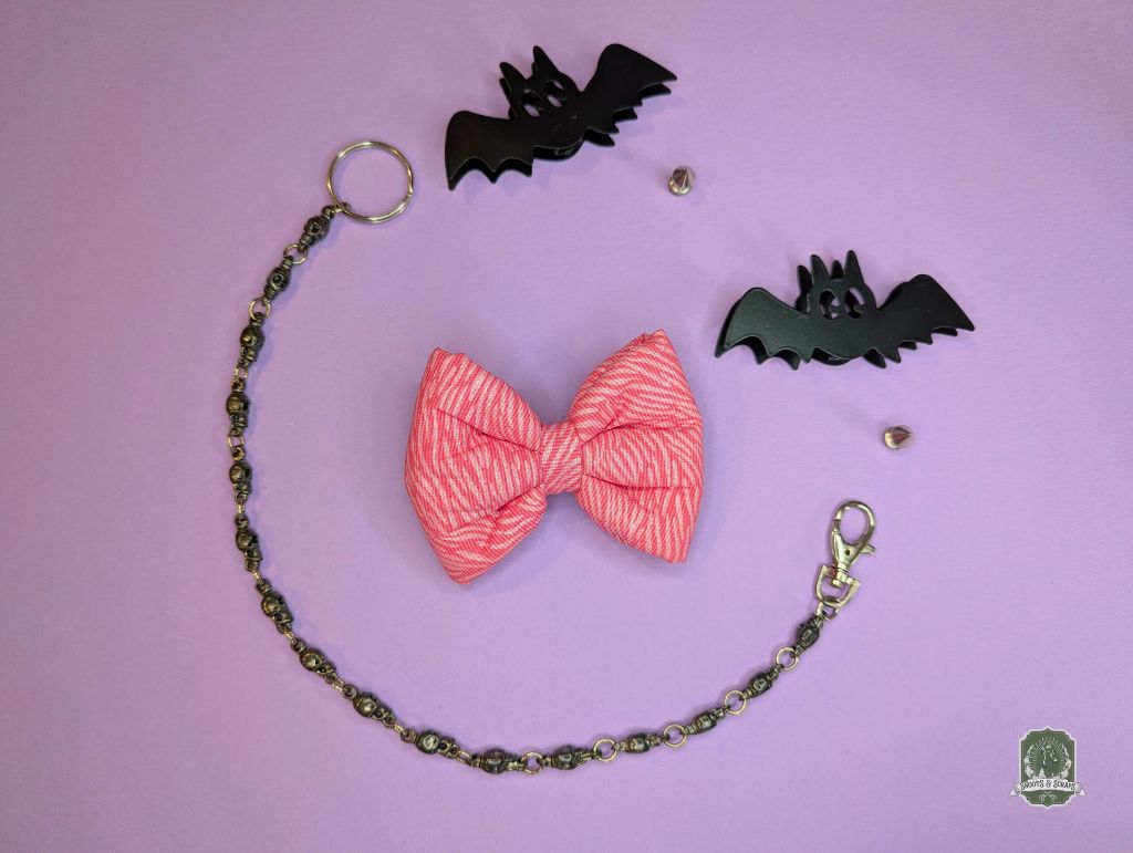Pink zebra print pet bow ties displayed on a pastel pink background and decorative chain and bat figurines.