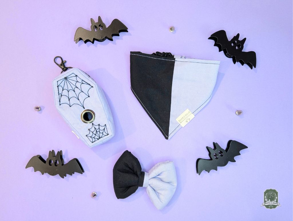 Halloween-themed pet accessories including a black and lilac bow tie,  black and lilac elastic dog bandana and coffin shaped poop bag holder on a purple background.