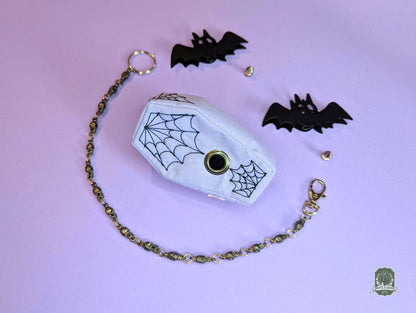 lilac coffin shaped poop bag holder with black embroidered spiderwebs on a pastel pink background
