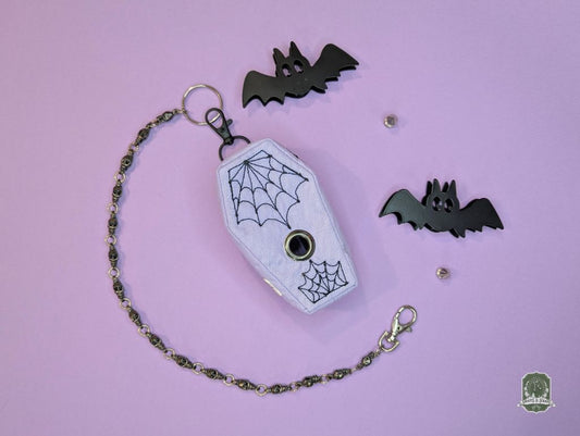 lilac coffin shaped poop bag holder with black embroidered spiderwebs on a pastel pink background