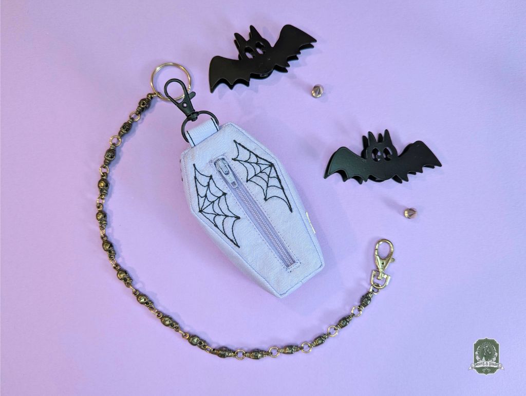 lilac coffin shaped poop bag holder with black embroidered spiderwebs on a pastel pink background