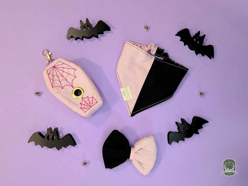 Halloween-themed accessories including a pink coffin shaped poo bag dispenser with spider web design, black and pink bandana, and a matching bow on a purple background.